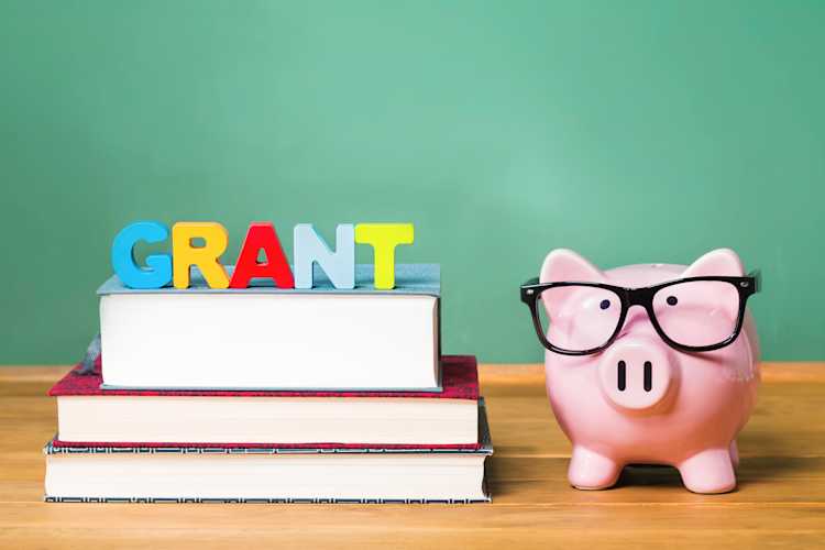 A wooden table displays a piggy bank and books, with a green board in the background, illustrating K-12 school safety grants. A wooden table displays a piggy bank and books, with a green board in the background, illustrating K-12 school safety grants.