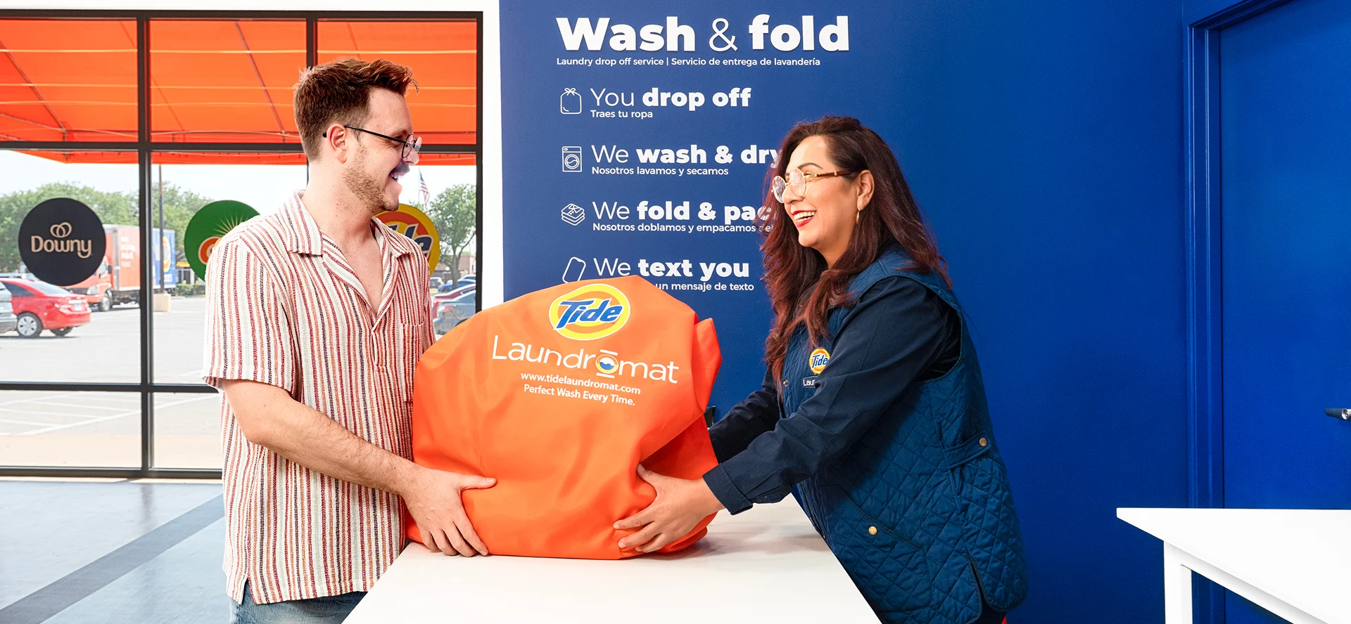 Cliente y empleado de Lavandería Tide intercambiando una bolsa de lavado y doblado en el mostrador.