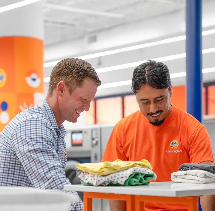 Store owner and Tide Laundromat employee smiling while folding freshly dried clothes inside the laundromat.