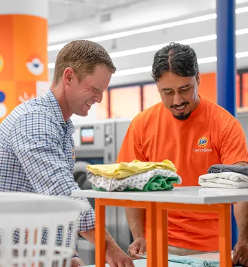 Store owner and Tide Laundromat employee smiling while folding freshly dried clothes inside the laundromat.