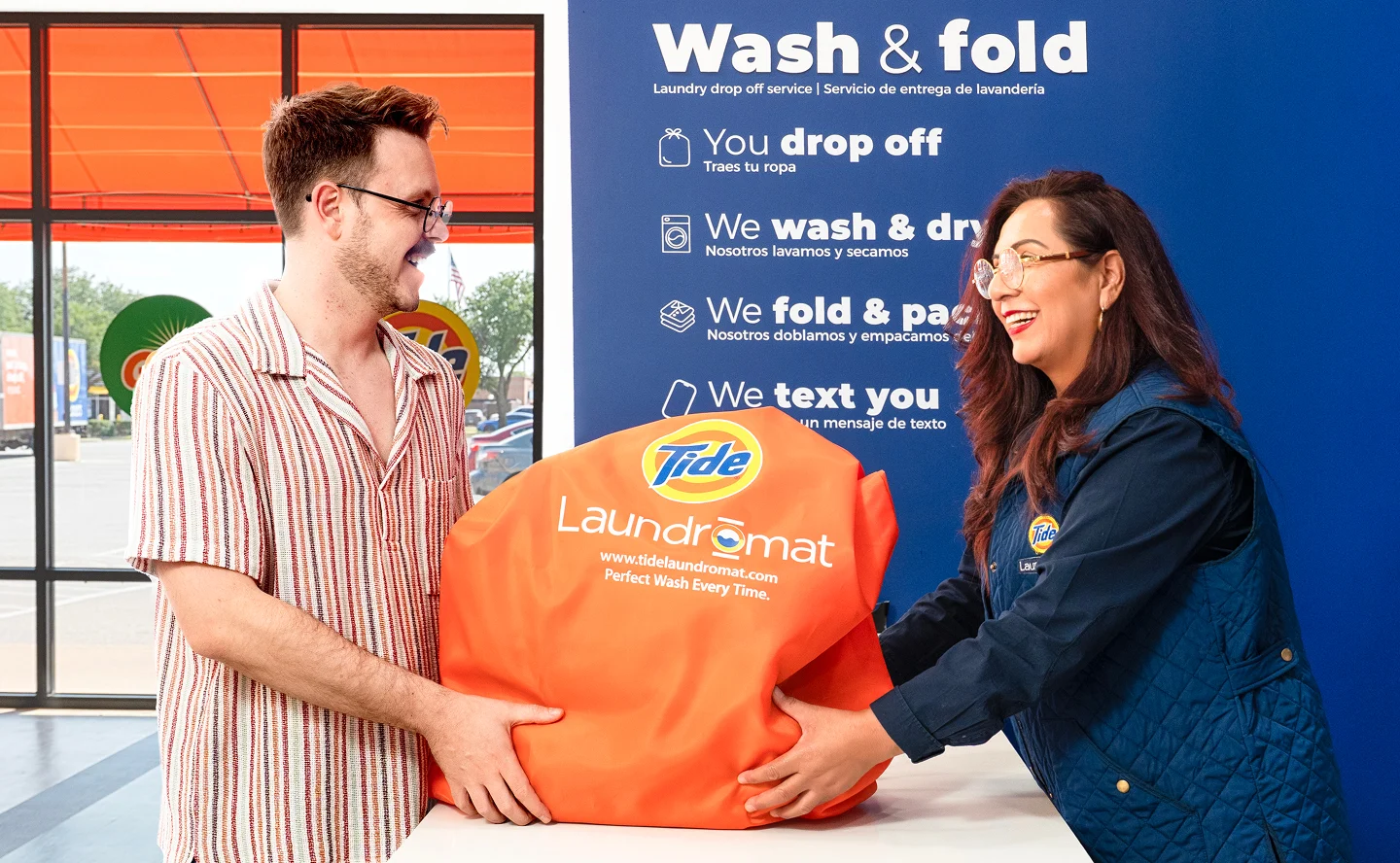 Cliente y empleado de Lavandería Tide intercambiando una bolsa de lavado y doblado en el mostrador.