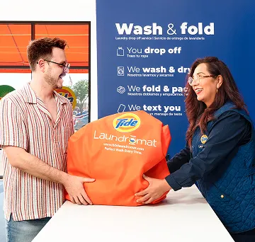 Cliente y empleado de Lavandería Tide intercambiando una bolsa de lavado y doblado en el mostrador.