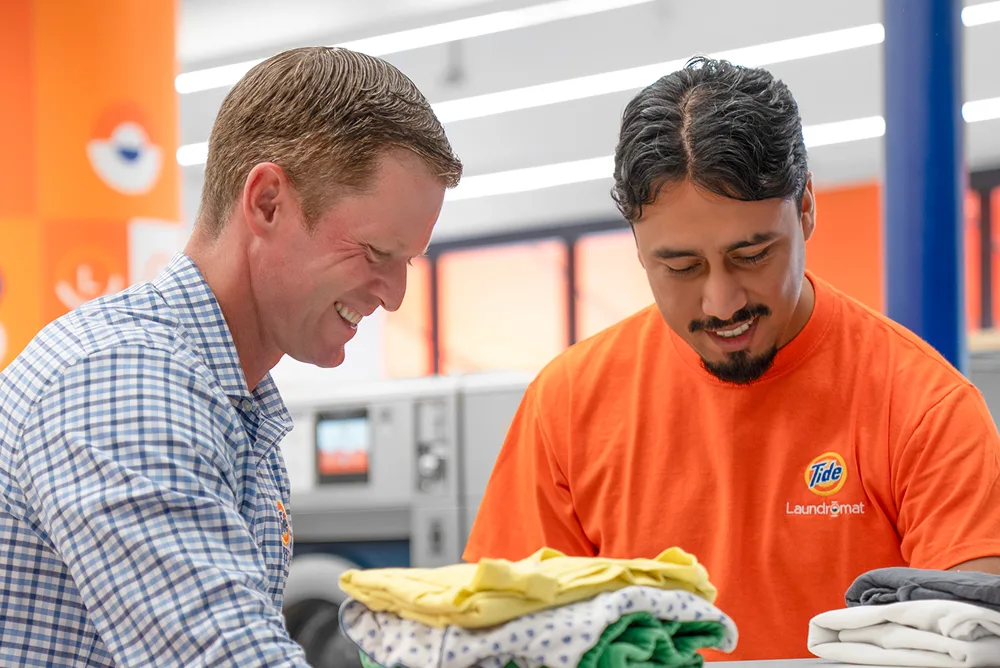 Store owner and Tide Laundromat employee smiling while folding freshly dried clothes inside the laundromat.
