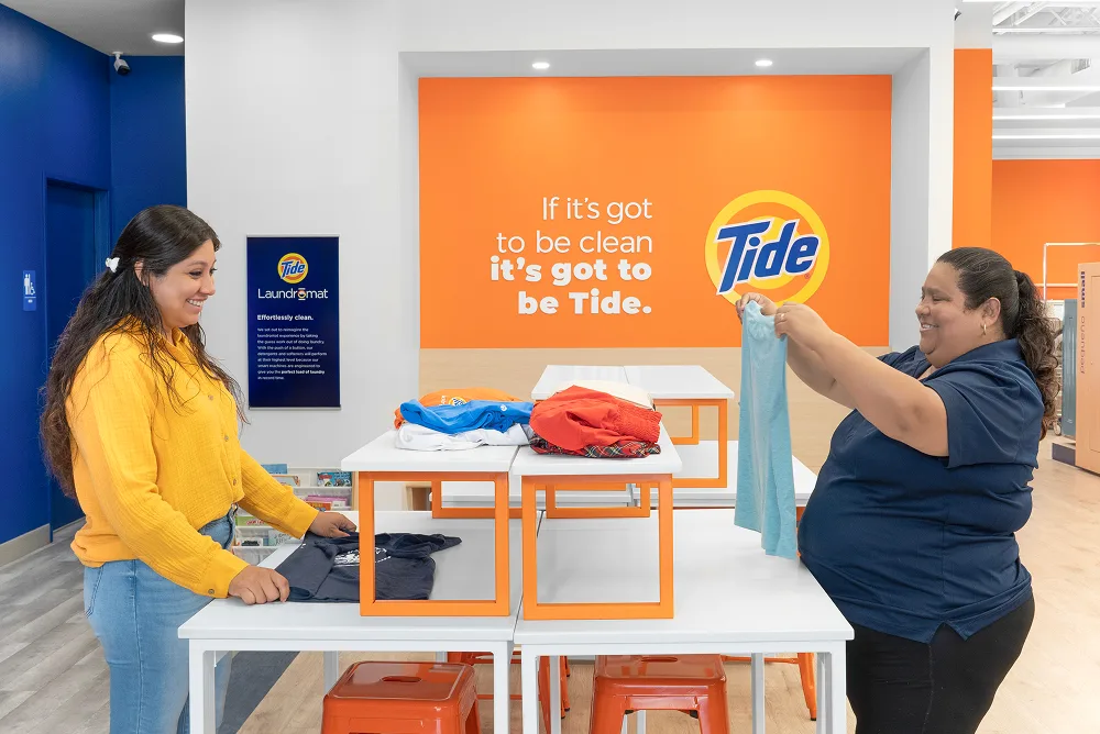 Store manager and customer smiling while folding clean laundry inside a laundromat.