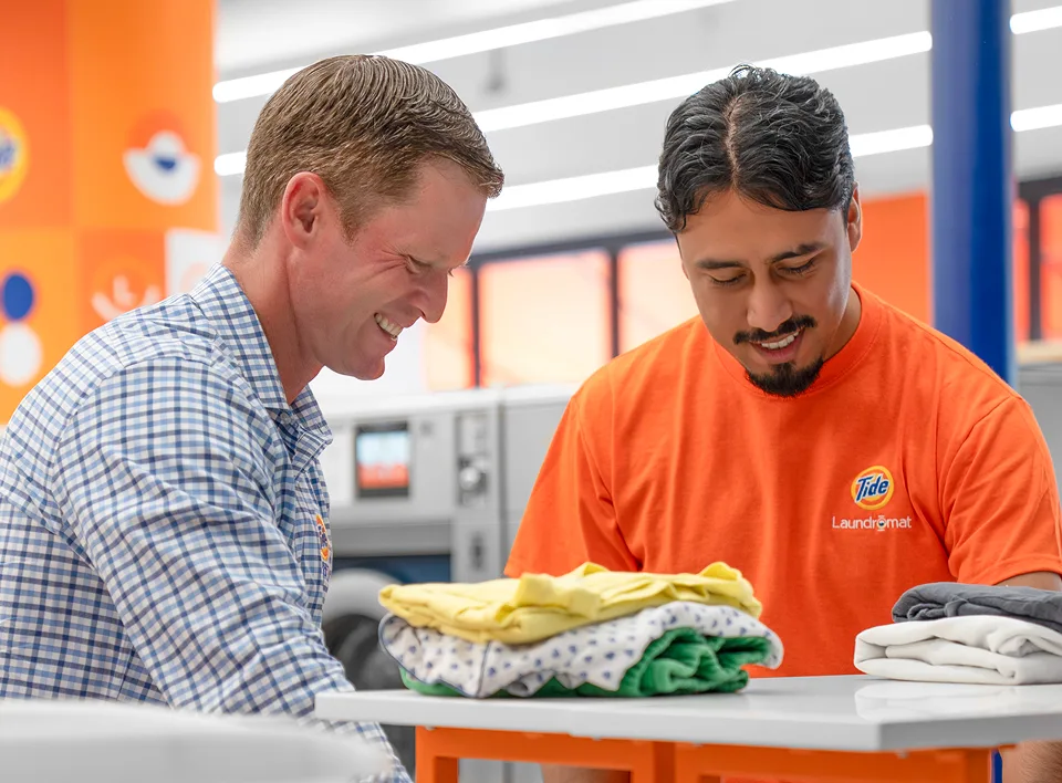 Store owner and Tide Laundromat employee smiling while folding freshly dried clothes inside the laundromat.