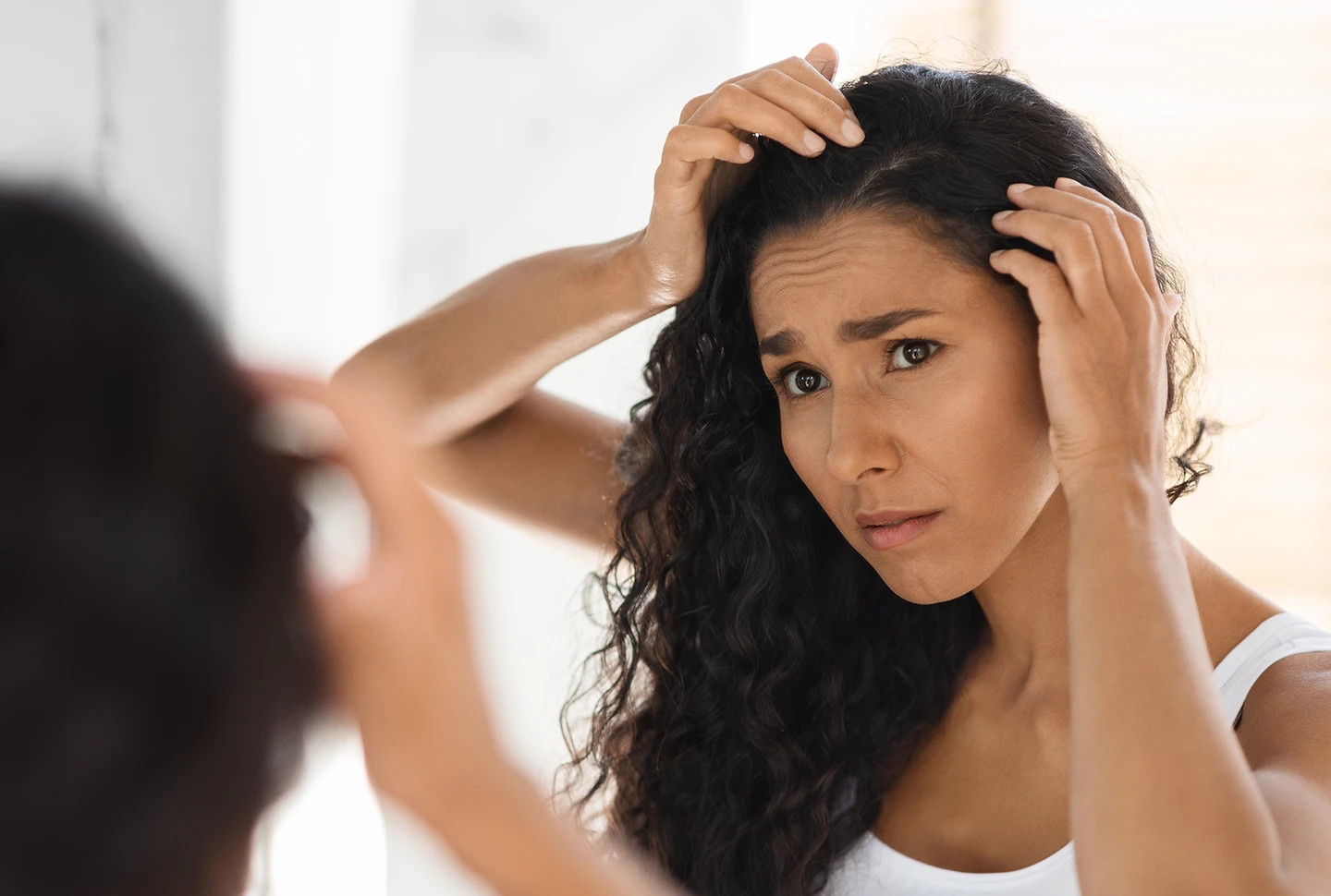A tan-skinned woman with long curly black hair and dry scalp looking into mirror to check dandruff with distressed look. A tan-skinned woman with long curly black hair and dry scalp looking into mirror to check dandruff with distressed look.