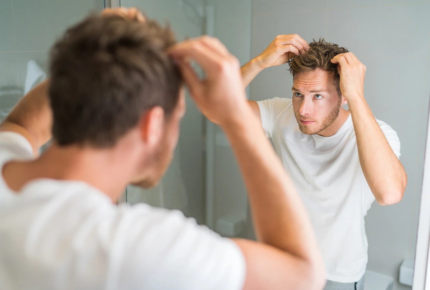 A fair-skinned man with oily scalp looks into mirror with hands on his head inspecting any dandruff in his brown hair in a bathroom. A fair-skinned man with oily scalp looks into mirror with hands on his head inspecting any dandruff in his brown hair in a bathroom.