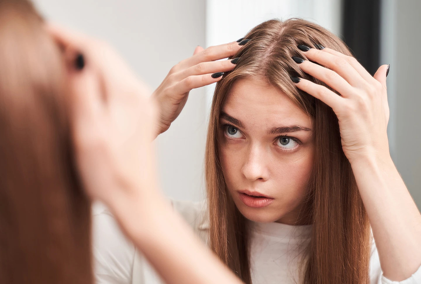 A fair-skinned girl with long strawberry blonde hair wearing a white t-shirt inspects her scalp in a mirror. A fair-skinned girl with long strawberry blonde hair wearing a white t-shirt inspects her scalp in a mirror.