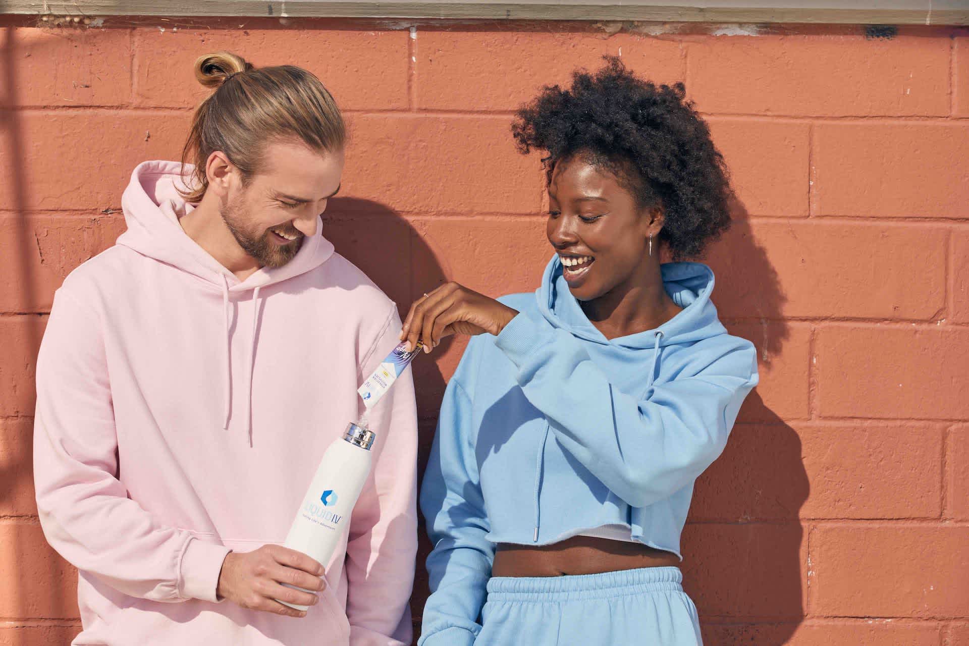 A young woman and a young man are standing against a brick wall. She is pouring a stick of Kygo Piña Colada Hydration Multiplier into his steel water bottle. 