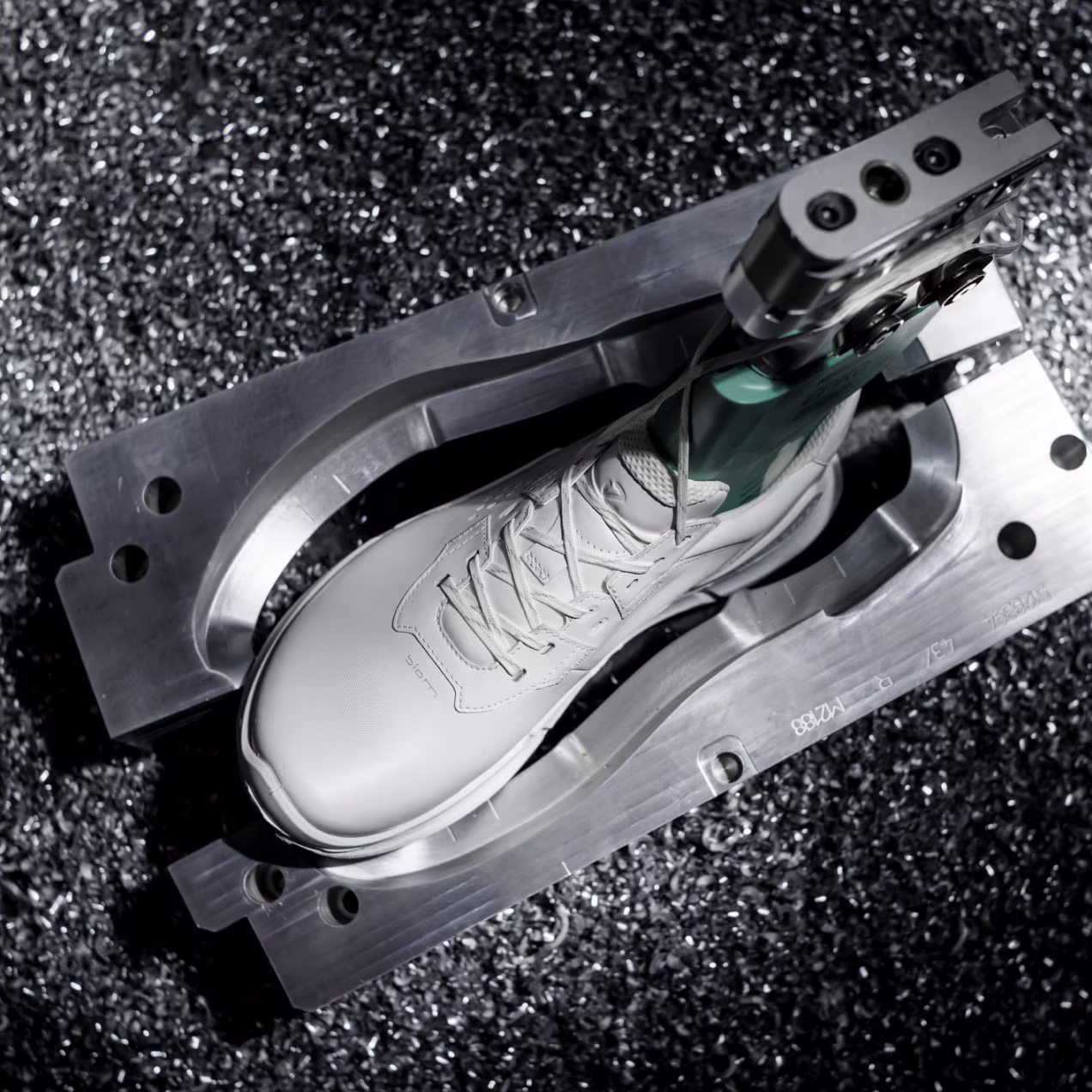 Close up look from above at the ECCO FLUIDFORM manufacturing process, showing a white sneaker secured inside an open metal mold surrounded by industrial metal shavings.
