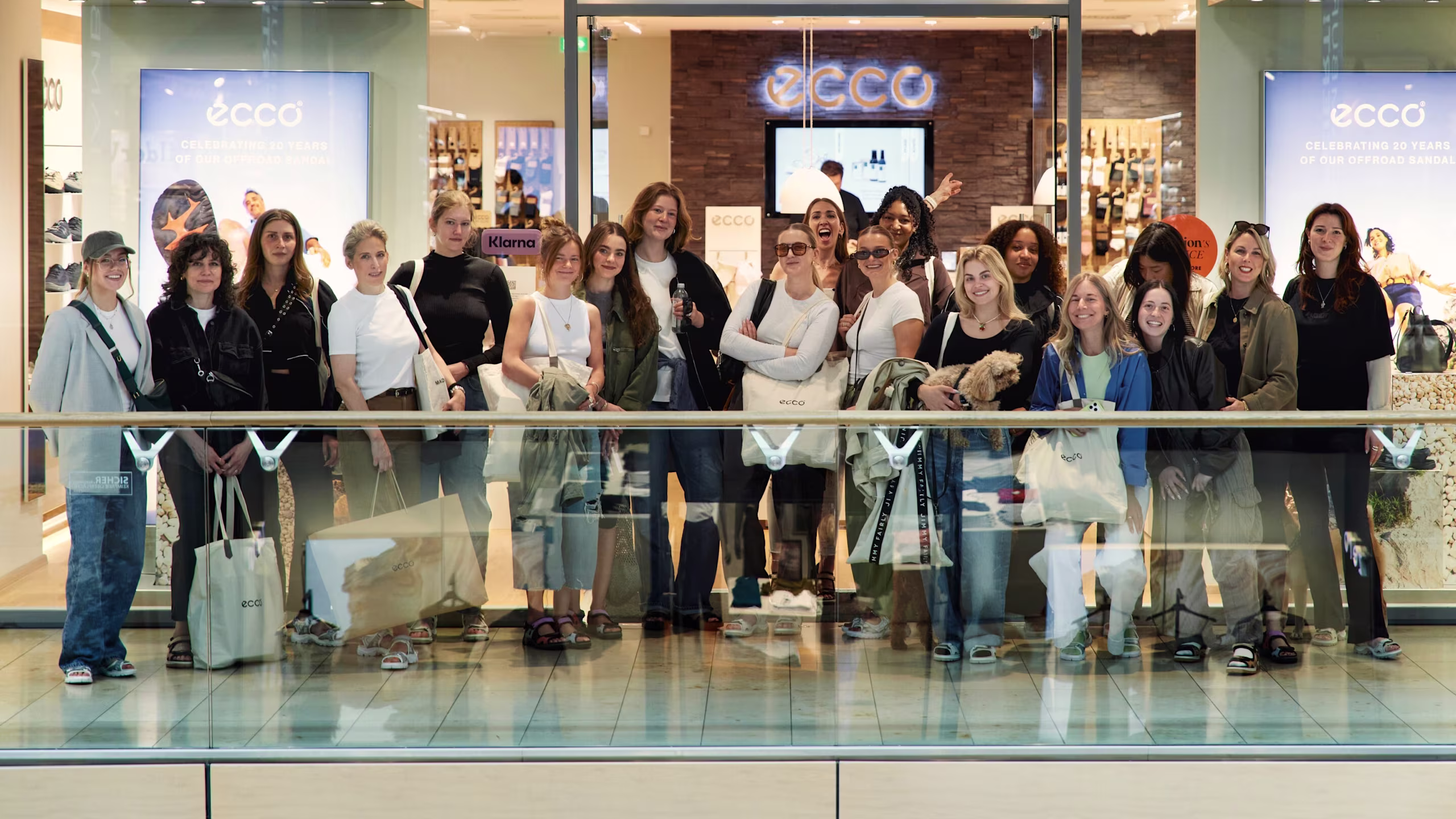 Eine fröhliche Gruppe in verschiedenen ECCO-Schuhen und mit ECCO-Einkaufstaschen versammelt sich vor der modernen Glasfront eines ECCO-Stores in einem belebten Einkaufszentrum.