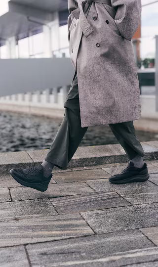 Persona caminando al aire libre con zapatillas negras ECCO, calcetines grises, pantalón oscuro y gabardina beis
