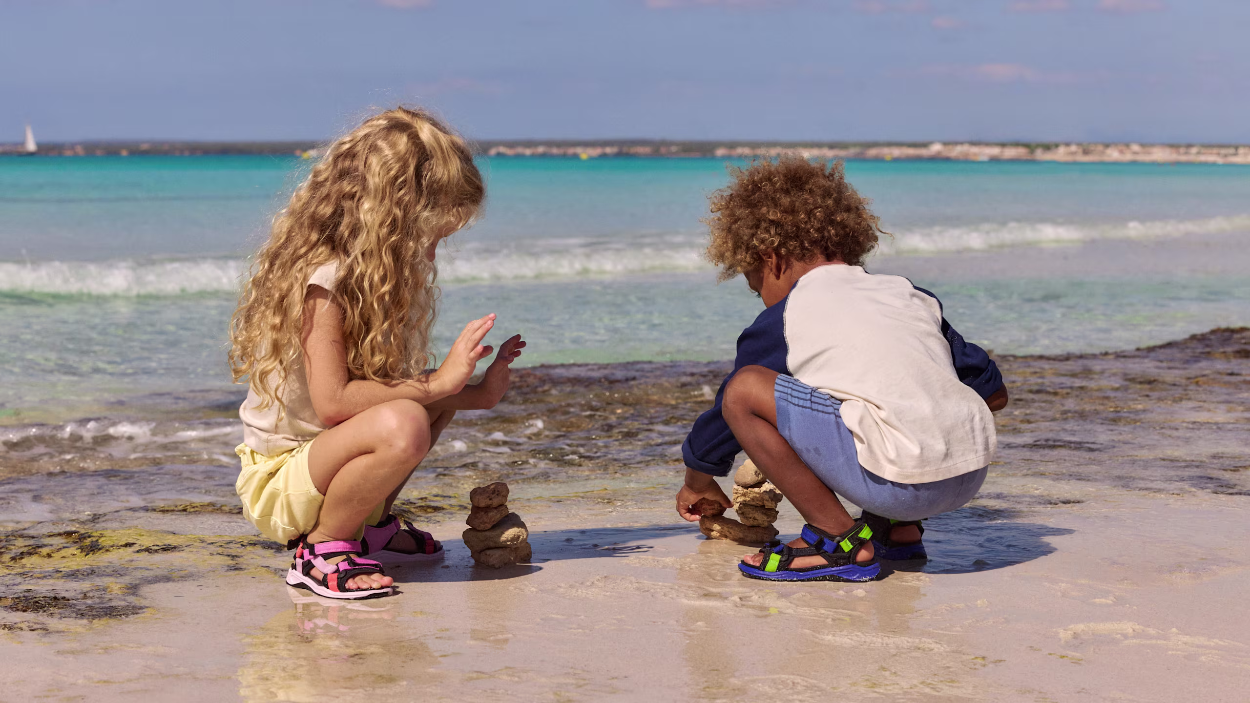 Kids in active ECCO multi-strap sandals crouching to play with rocks on a sunny beach.