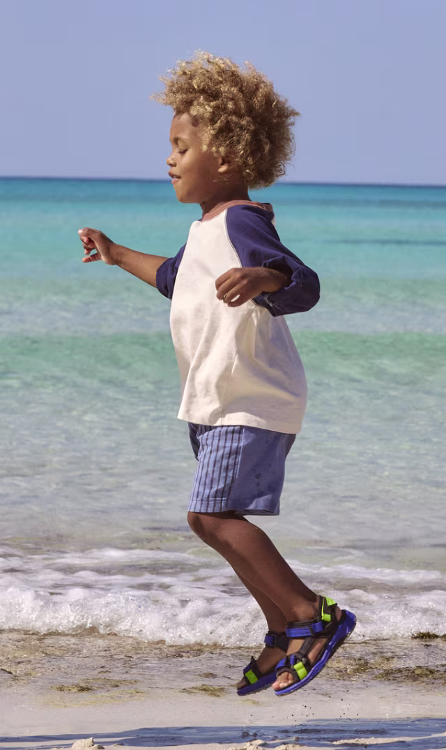 ECCO X-Trinsic kids' sporty blue and green sandals, worn by a child jumping on a sandy beach shoreline.