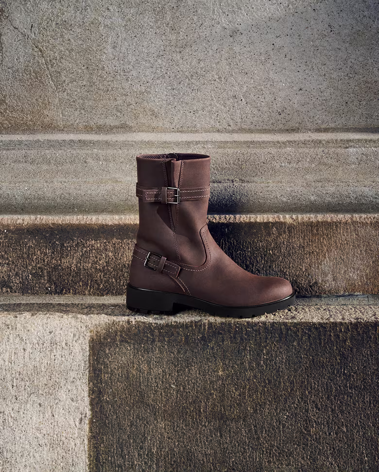 Ein Mann und eine Frau in eleganten Mänteln neben einem braunen, halbhohen Lederstiefel mit Schnalle.