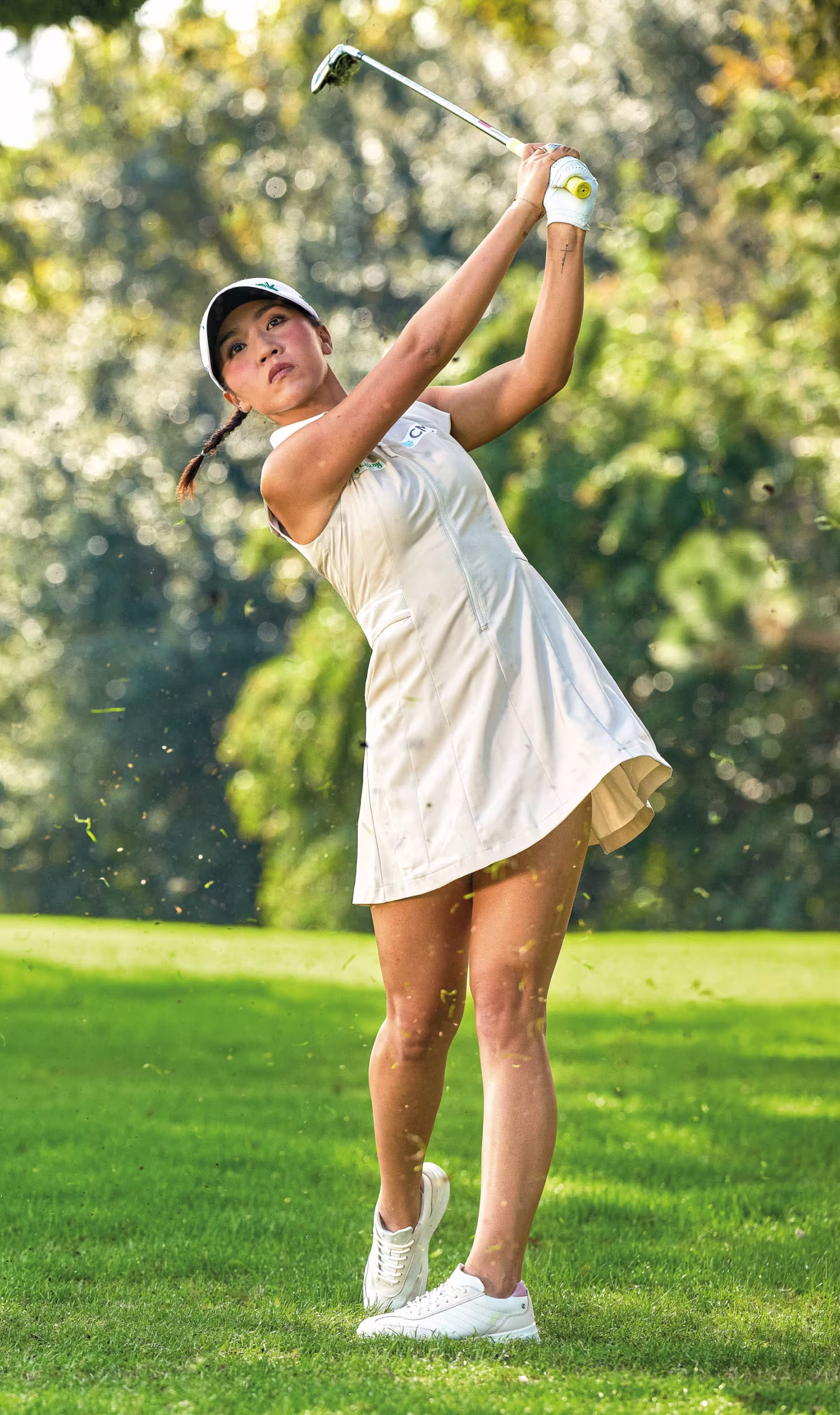 Golfista a meio da tacada, usando sapatos de golfe ECCO em pele branca e vestido de golfe creme sem mangas num campo verde.