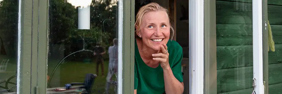 En leende person lutar sig mot ett öppet fönster i ett grönt trähus med en telefon i handen. I bakgrunden syns en trädgård i sommarkvällens ljus.
