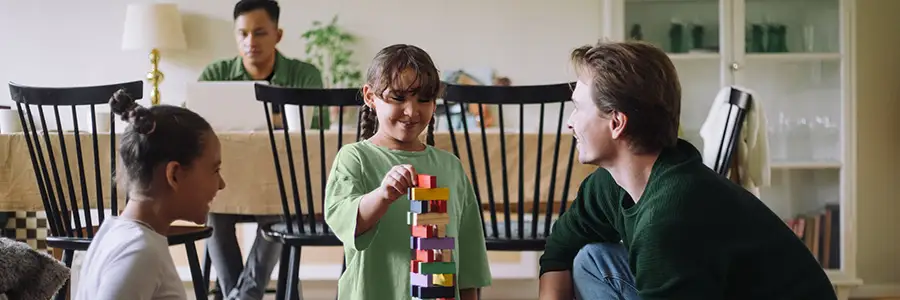 En familj tillbringar tid tillsammans i vardagsrummet. Två barn och en vuxen bygger ett torn av färgglada klossar medan en annan vuxen arbetar vid en bärbar dator vid bordet i bakgrunden.