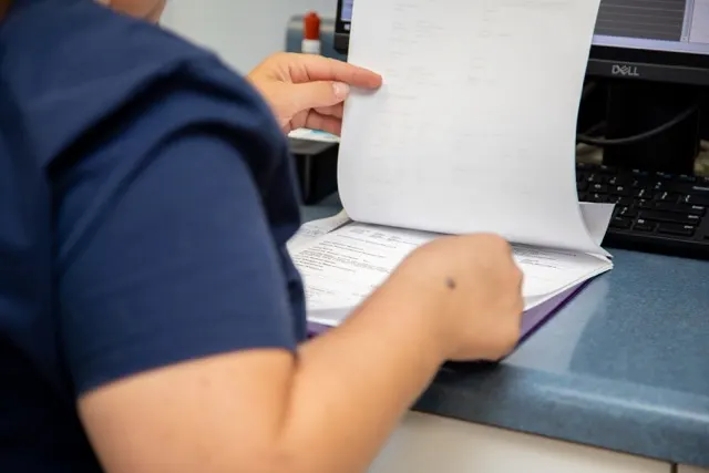 A close-up view of a veterinary professional in dark blue scrubs flipping through pages in a purple folder at a desk next to a computer. A close-up view of a veterinary professional in dark blue scrubs flipping through pages in a purple folder at a desk next to a computer.
