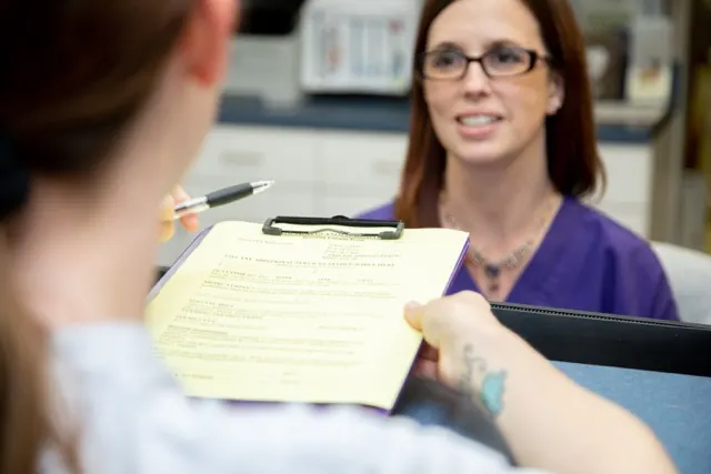 Over-the-shoulder view of a client handing a yellow intake form on a clipboard to a veterinary receptionist wearing purple scrubs. Over-the-shoulder view of a client handing a yellow intake form on a clipboard to a veterinary receptionist wearing purple scrubs.
