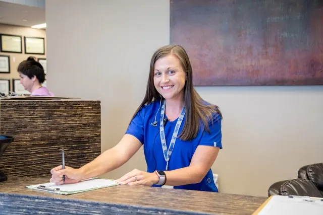 A smiling veterinary receptionist in blue scrubs sitting at a front desk, writing on a clipboard.