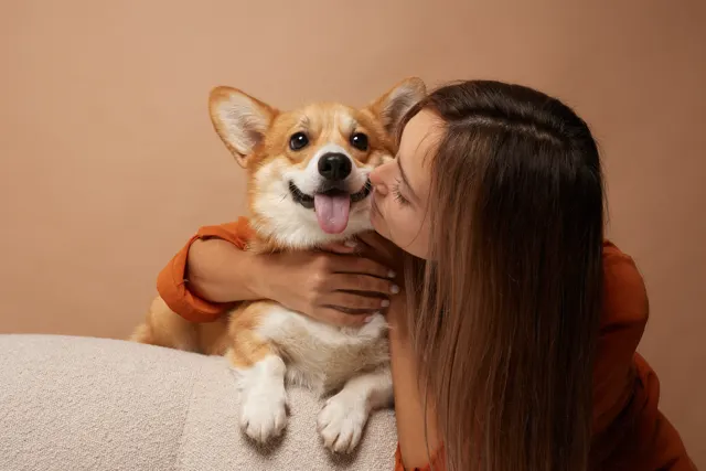 A woman with long brown hair leaning in to kiss a Pembroke Welsh Corgi on the cheek; the Corgi is smiling with its tongue out, and both are positioned against a warm, tan background.