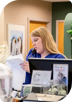 A busy receptionist talking on a landline phone while reviewing a stack of documents at a desk with a monitor and a donation jar.