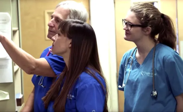 Three medical professionals (two female, one male) in blue scrubs reviewing patient documents on a clipboard or wall chart.