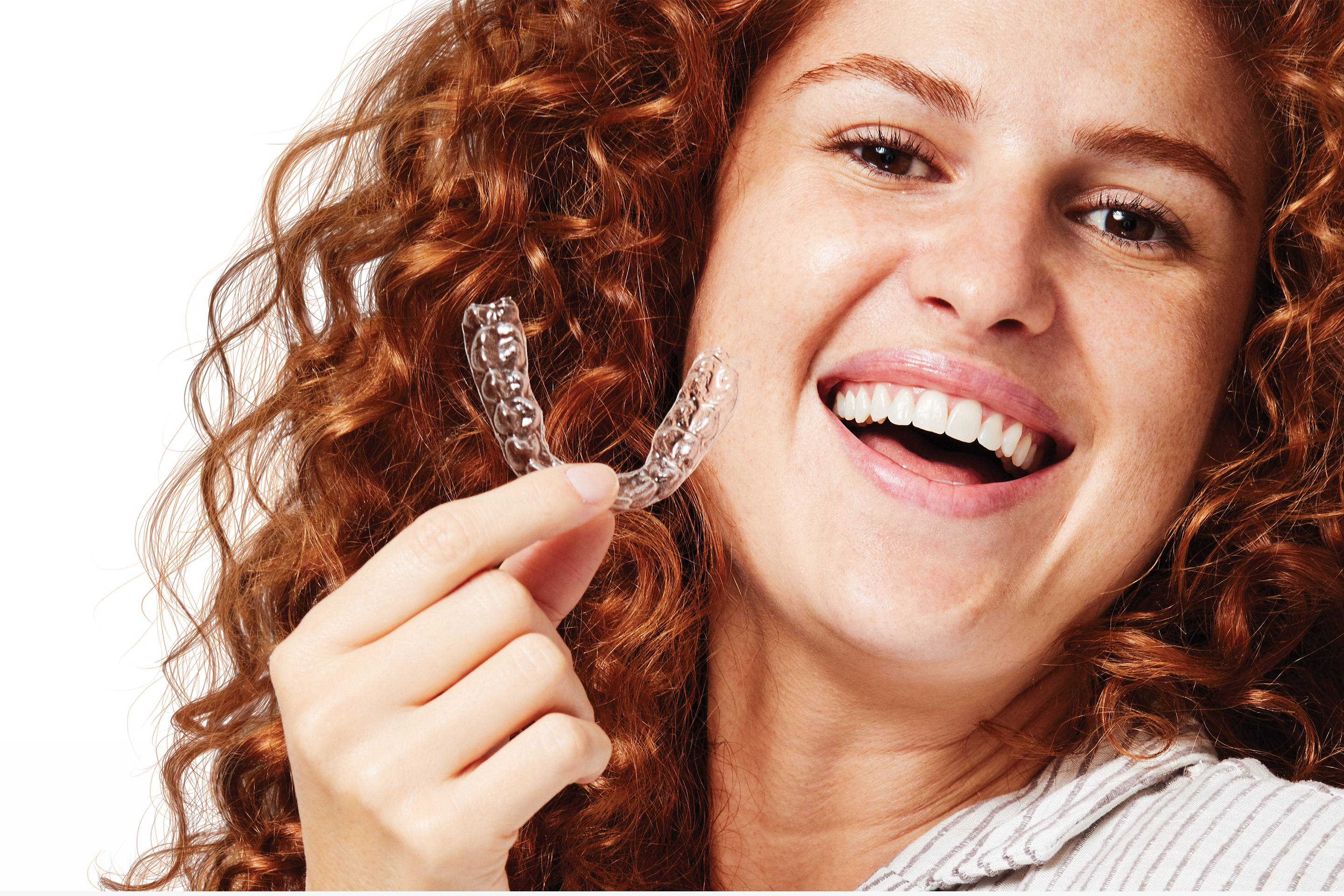 Close up headshot of a smiling woman with red curly hair holding SmileDirectClub aligner near her mouth