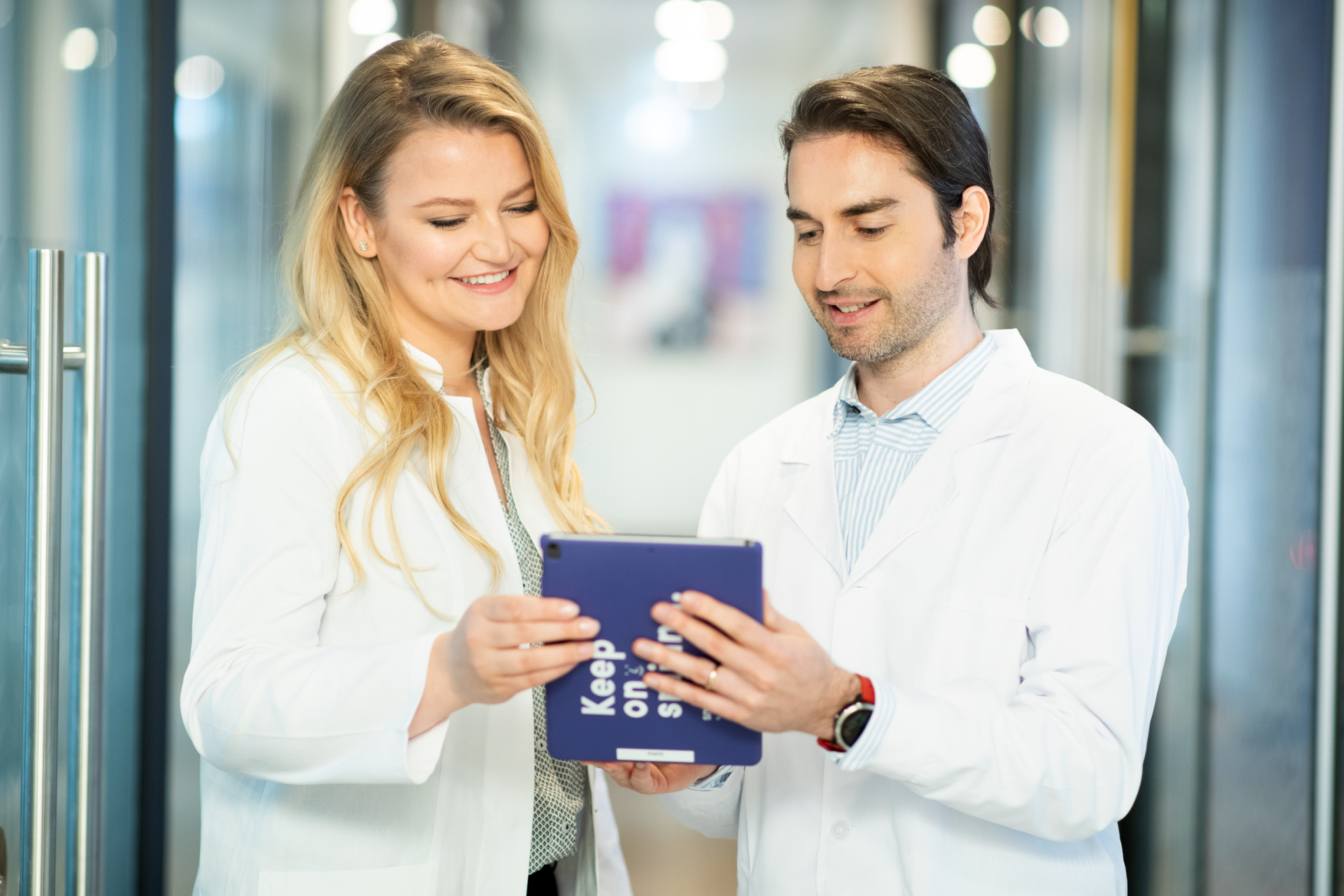 A blonde woman and brunette man wearing lab coats smiling and looking at a SmileDirectClub tablet