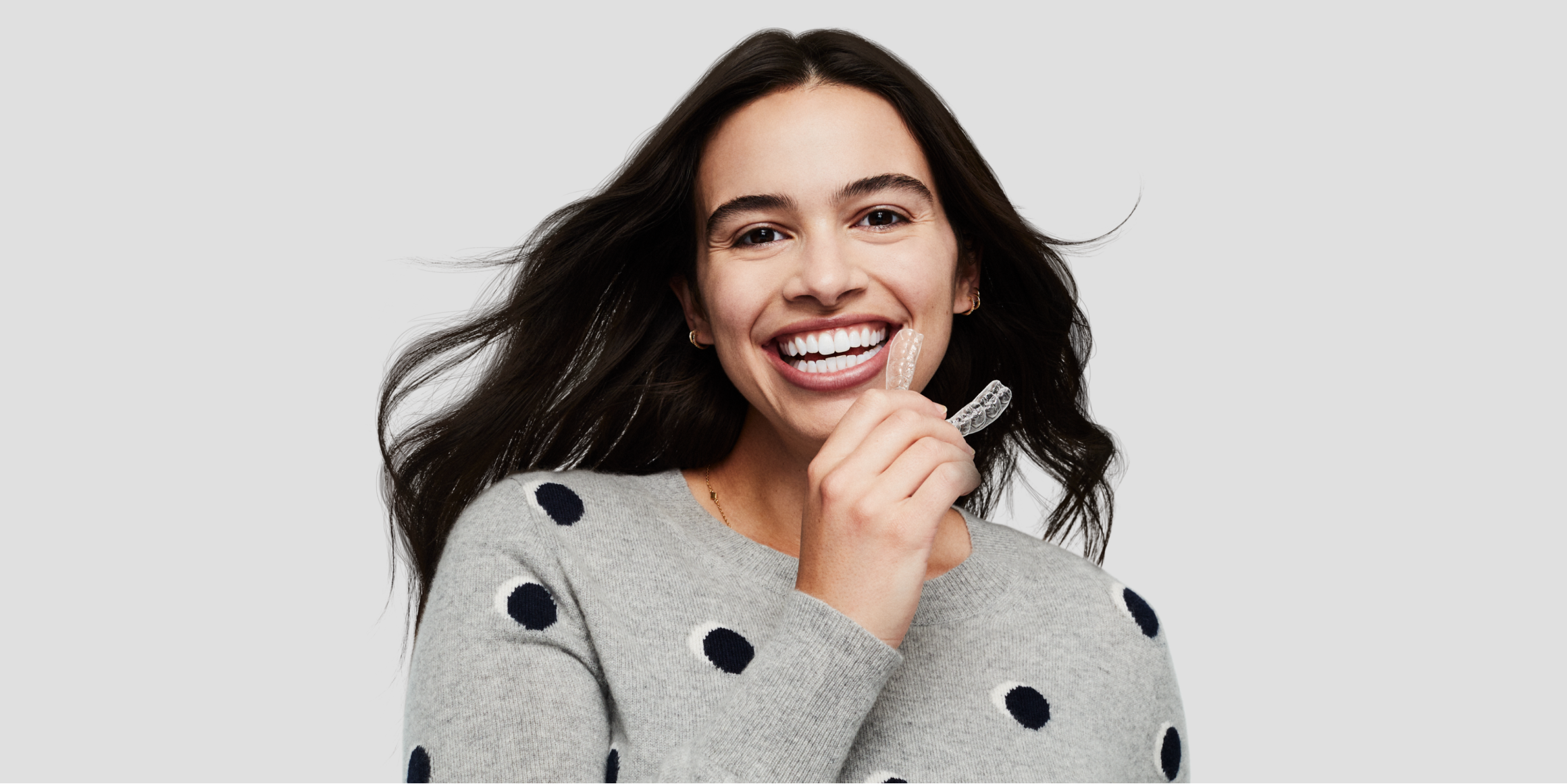 A brunette woman in a gray sweater smiling and holding a smiledirectclub clear aligner near her mouth 