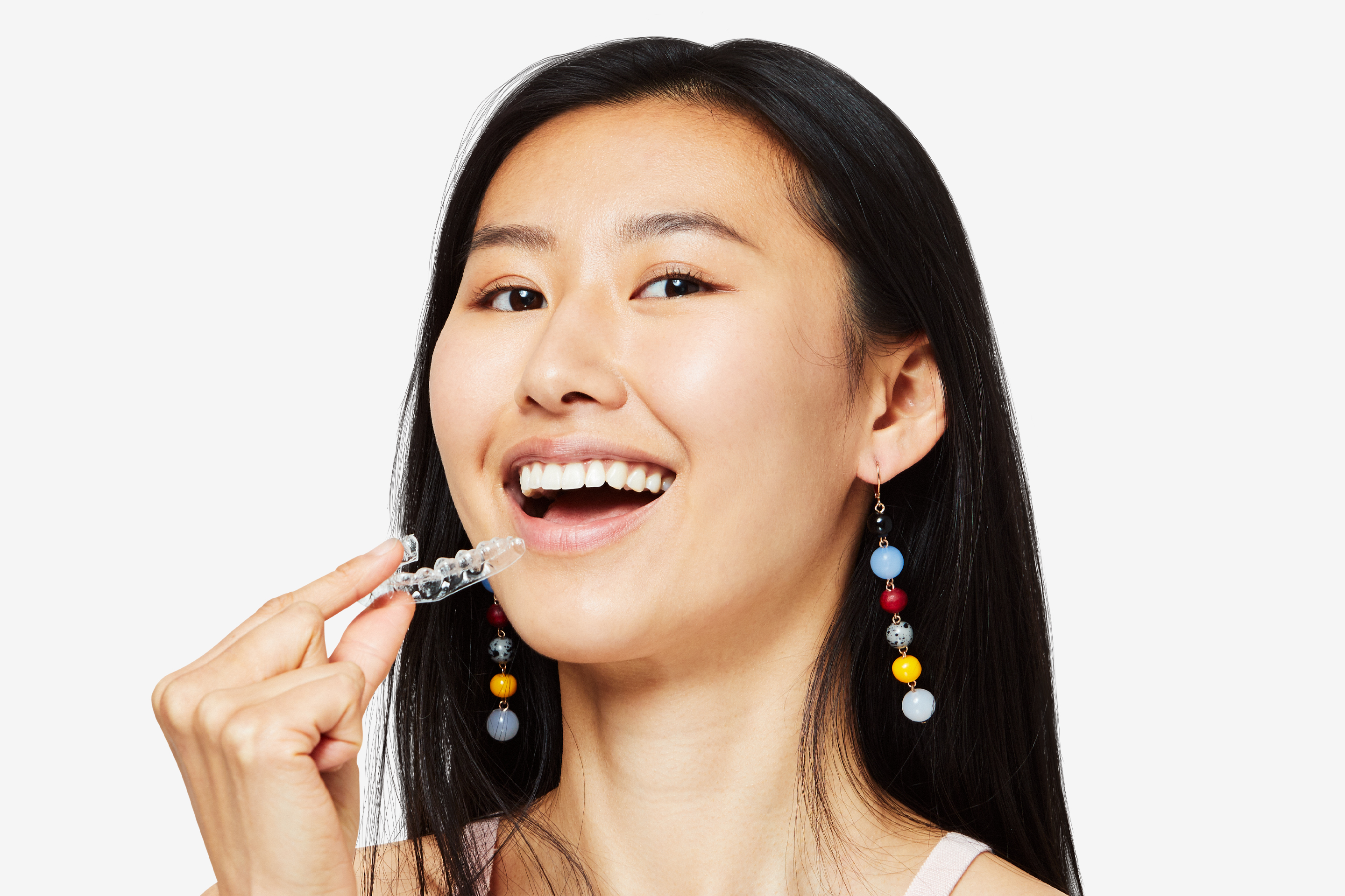 Asian woman smiling with colorful earrings holding a SmileDirectClub clear aligner near her mouth