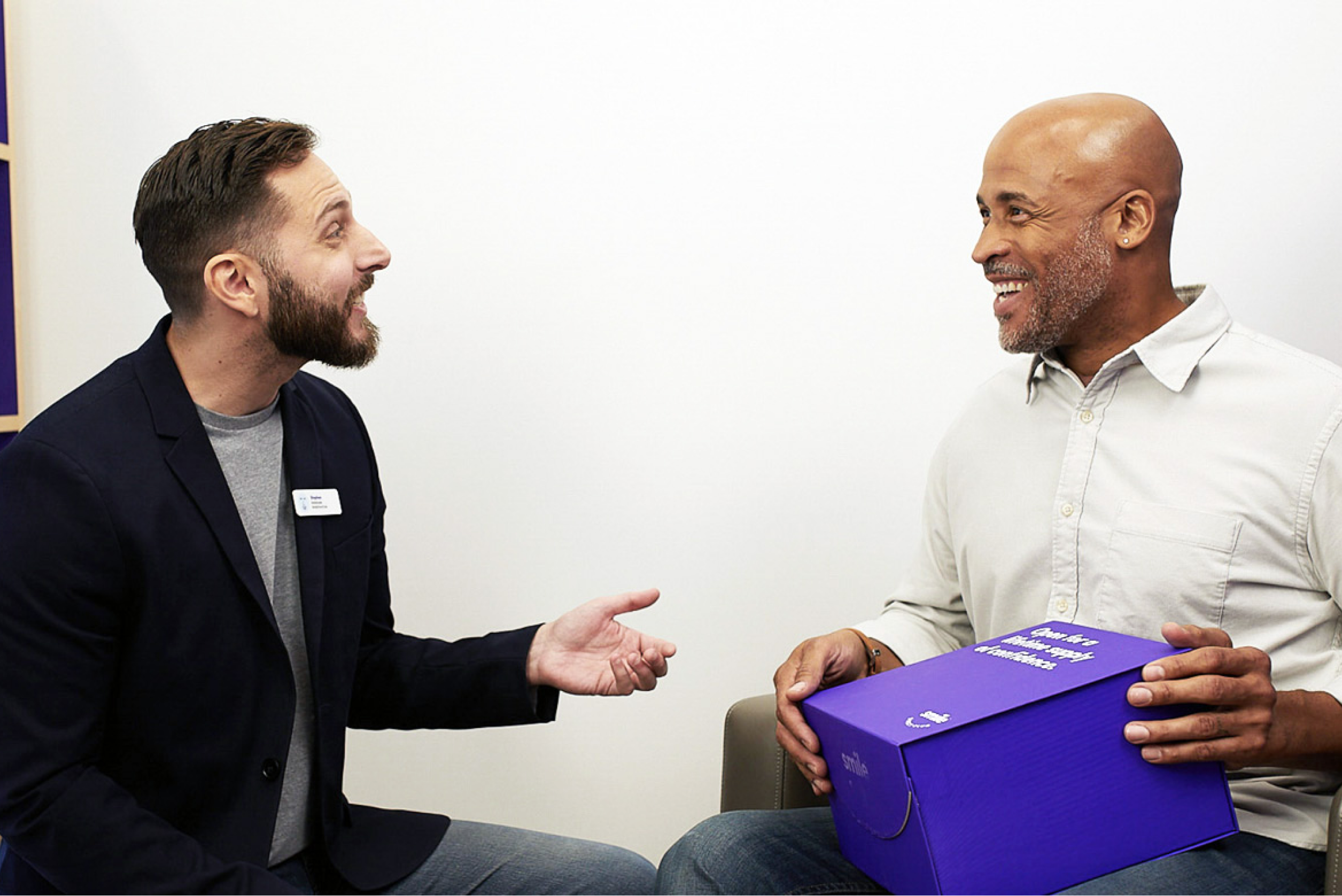 Man seated holding a Burple SmileDirectClub Aligner Box talking with SmileGuide at a Smileshop