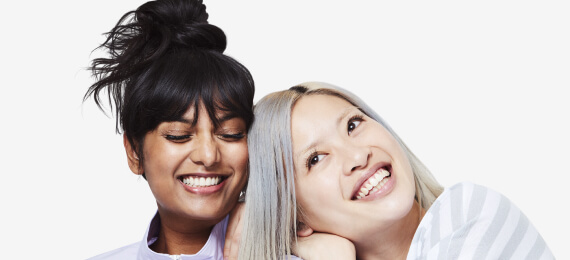 Blonde Asian woman lying on an Black woman's shoulder smiling for smiledirectclub