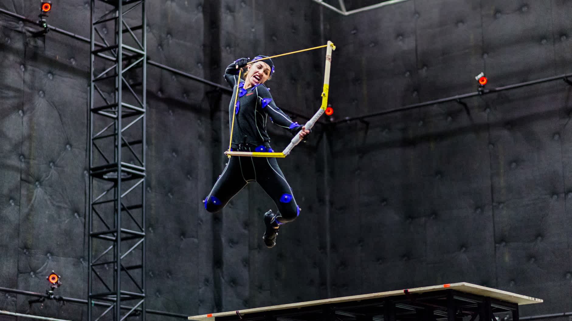 Stunt woman, wearing a motion capture suit, jumping in the air with a bow arrow.