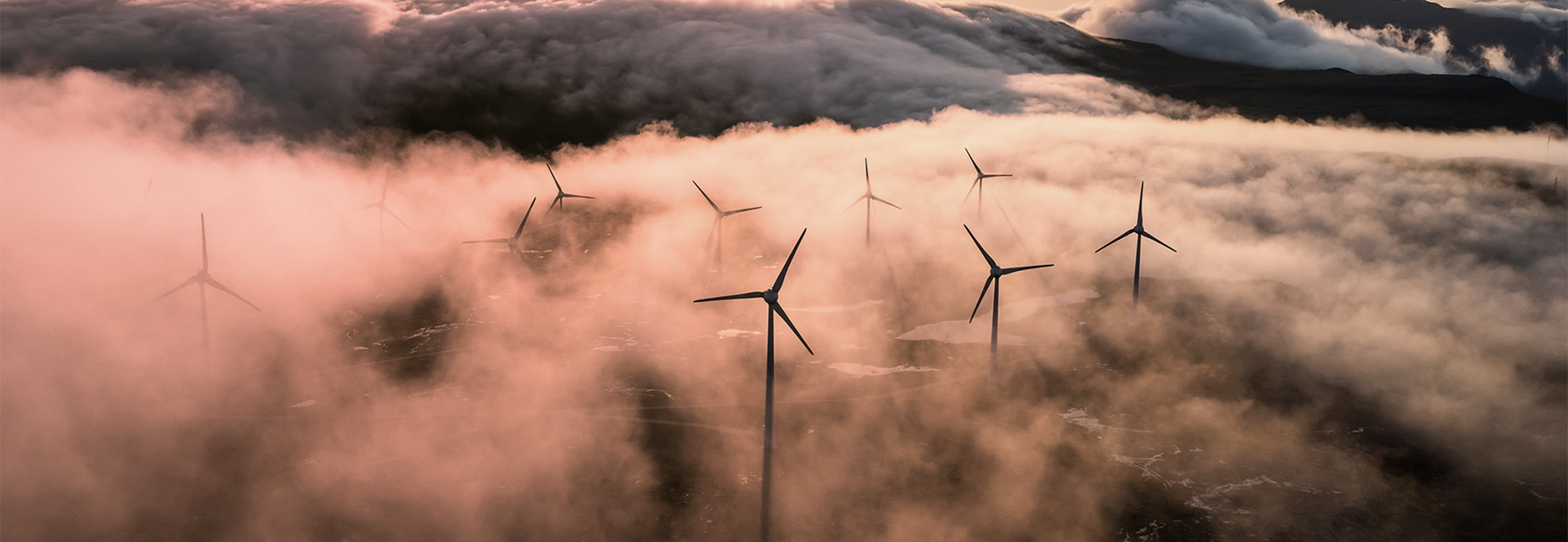 Wind turbines scattered across a mountainous landscape, surrounded by low-hanging mist and soft morning light.