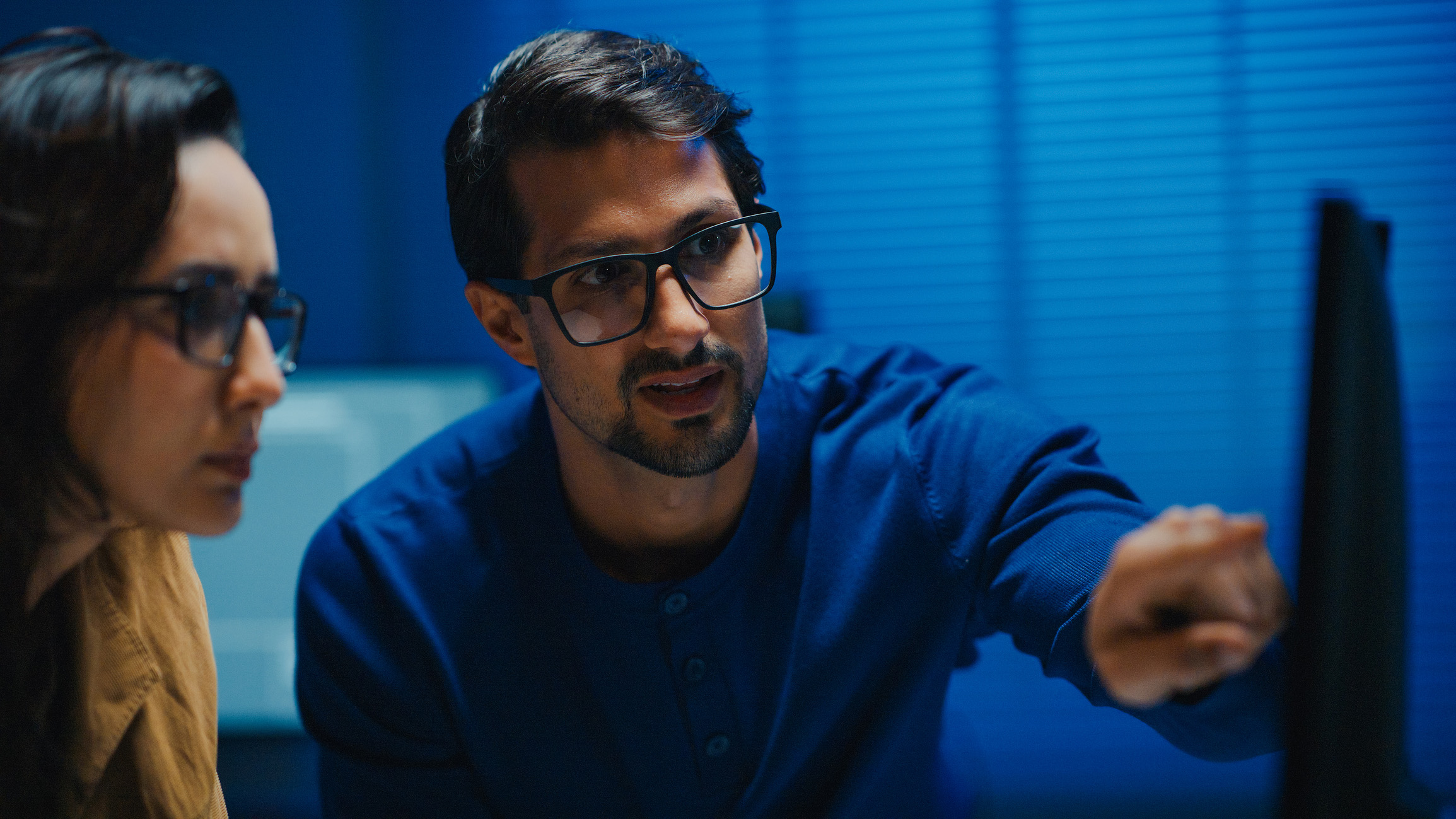 Two colleagues focused on an off-screen computer monitor. One of the colleague points toward the screen while working in a dark, blue-lit office.