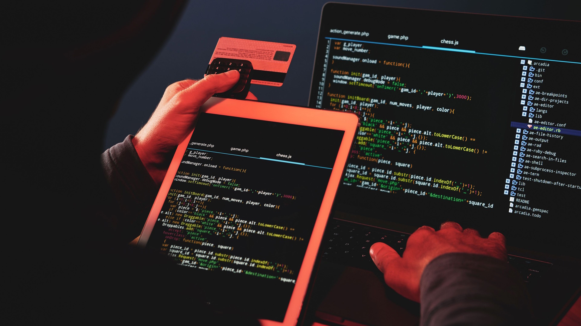 Person holding the back of a credit card and coding on laptop and tablet