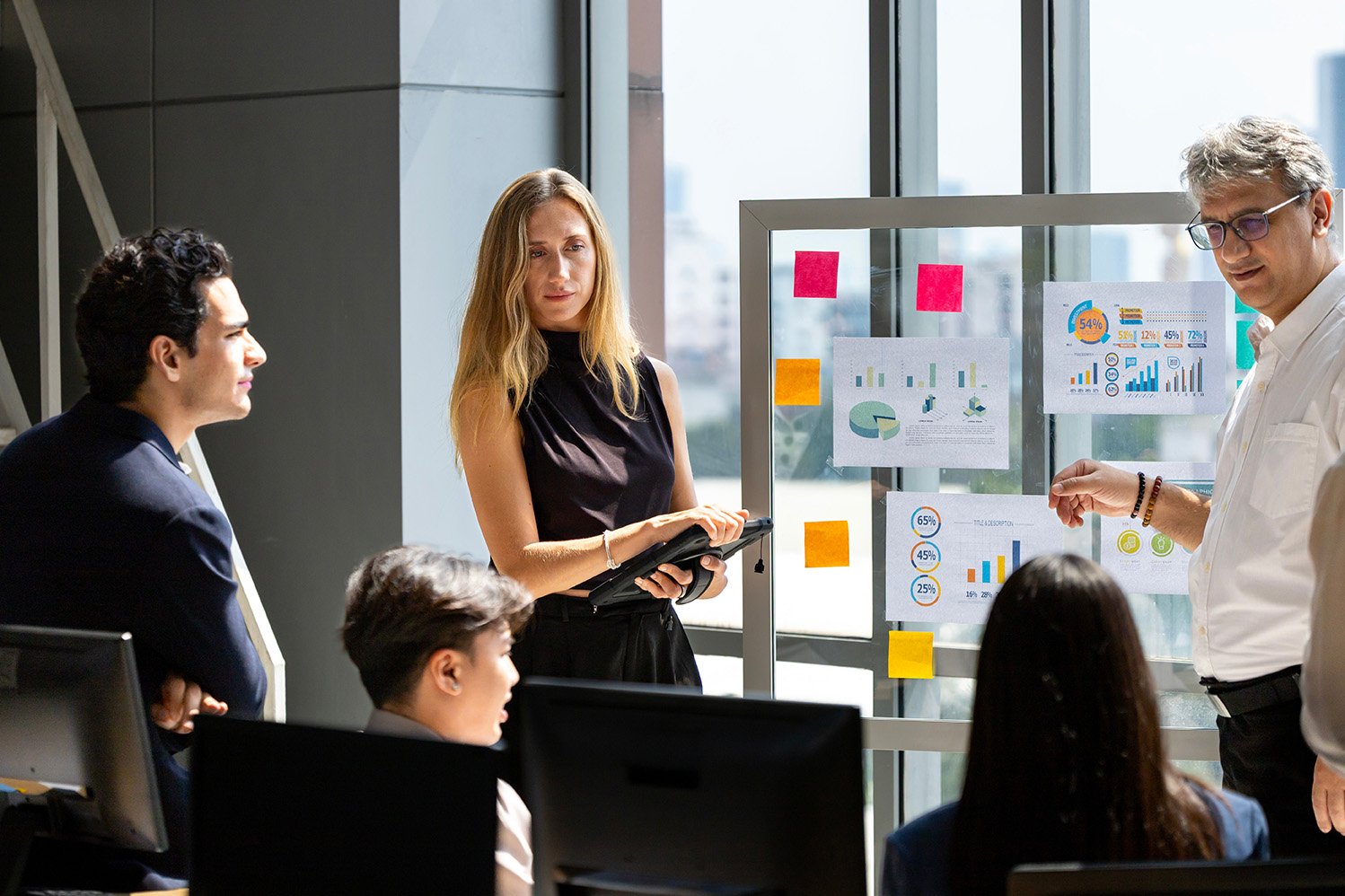 A man points to a glass whiteboard with charts and sticky notes, presenting to three other colleagues. One woman stands holding a tablet, while two others are seated, all listening in a bright office with large windows.