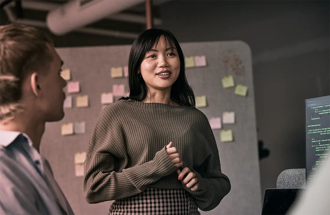 Young professional woman speaking during a team planning meeting with sticky notes and code on a monitor in the background.