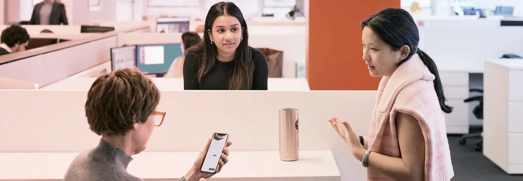 Three colleagues in a modern office speaking together, with one woman showing a smartphone screen while another gestures while talking.