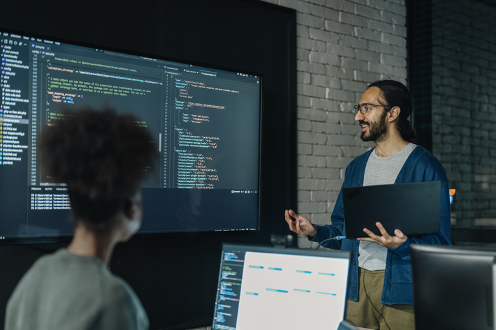 A man holds a laptop and gestures towards a large screen displaying code.