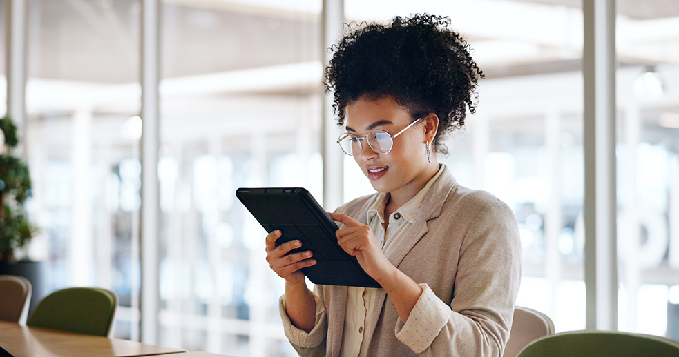 A woman with dark curly hair and glasses smiles as she holds and interacts with a tablet. She is standing in a modern, well-lit office with large windows and glass partitions in the background.