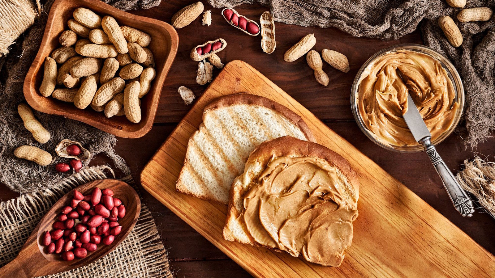 Two slices of bread, one with peanut butter, on the table with different peanuts around