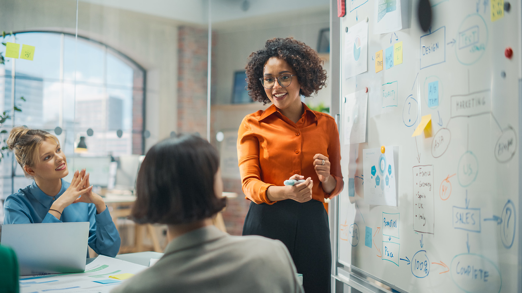 A smiling woman with curly hair stands at a whiteboard covered in diagrams and sticky notes, holding a marker. Two female colleagues are seated at a table watching her presentation in a bright office.
