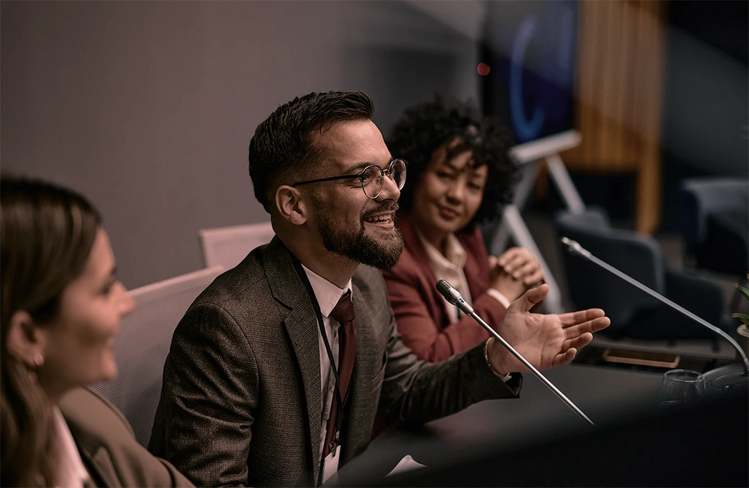 Man in a suit speaking into a microphone with hand gestures while sitting at a panel discussion table.