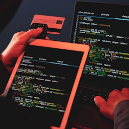 Thumbnail Person holding the back of a credit card and coding on laptop and tablet