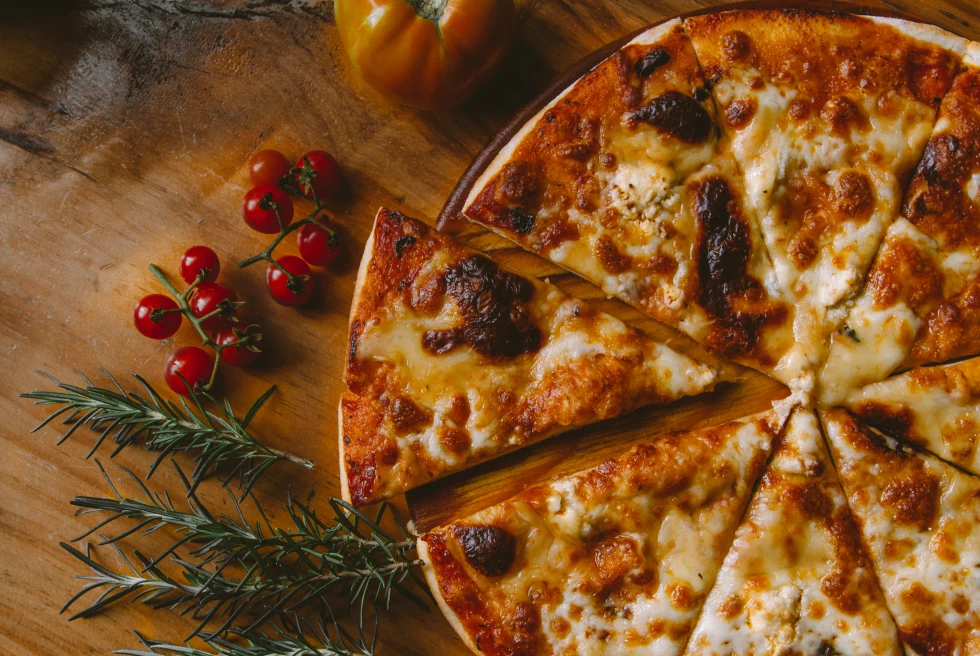 White and bubbly cheesy pizza with red sauce and thin crust on a wooden board with green rosemary and red tomatoes.