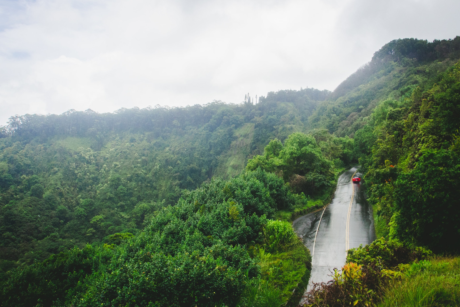 A pavement road with a red car driving up in the middle of lush greenery on The Road to Hana in Maui, Hawaii.