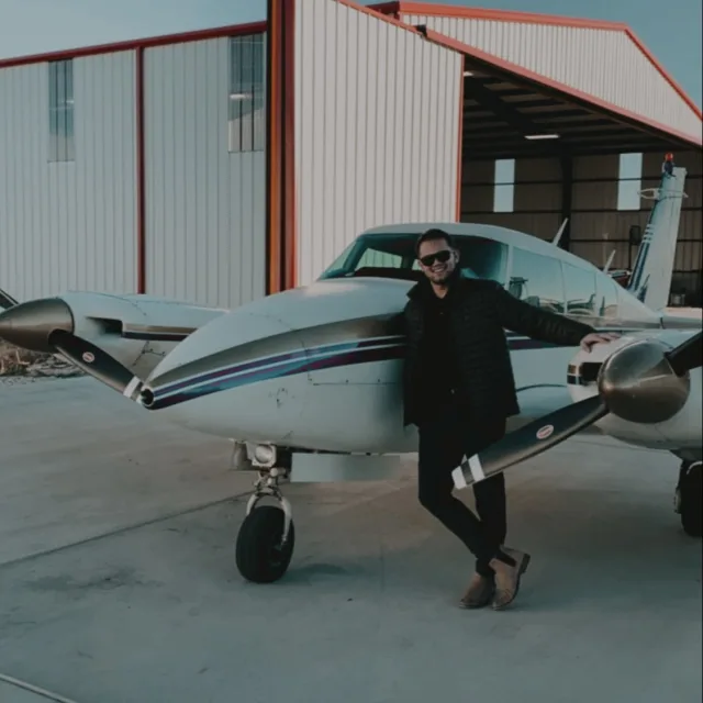 Dakk wearing a black jacket, dark pants and sunglasses while posing in front of a small airplane